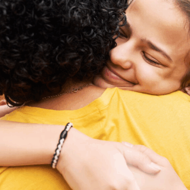 A woman hugs a friend. Her eyes are closed and she is smiling. The friend's back is to the camera.