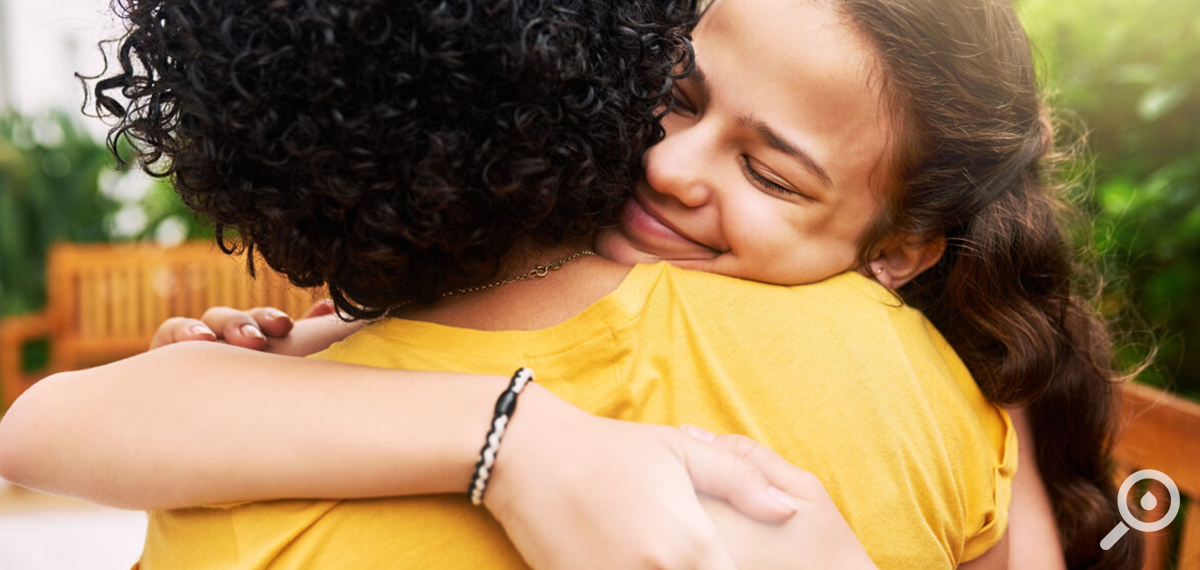 A woman hugs a friend. Her eyes are closed and she is smiling. The friend's back is to the camera.